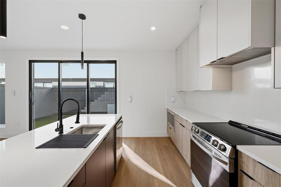 Kitchen features recessed lighting and pendant lighting over the island. Light wood cabinetry extends throughout with built-in microwave. Quartz waterfall island includes a black faucet and undermount sink. Sliding glass door provides access to the backyard patio. Vinyl flooring extends throughout.