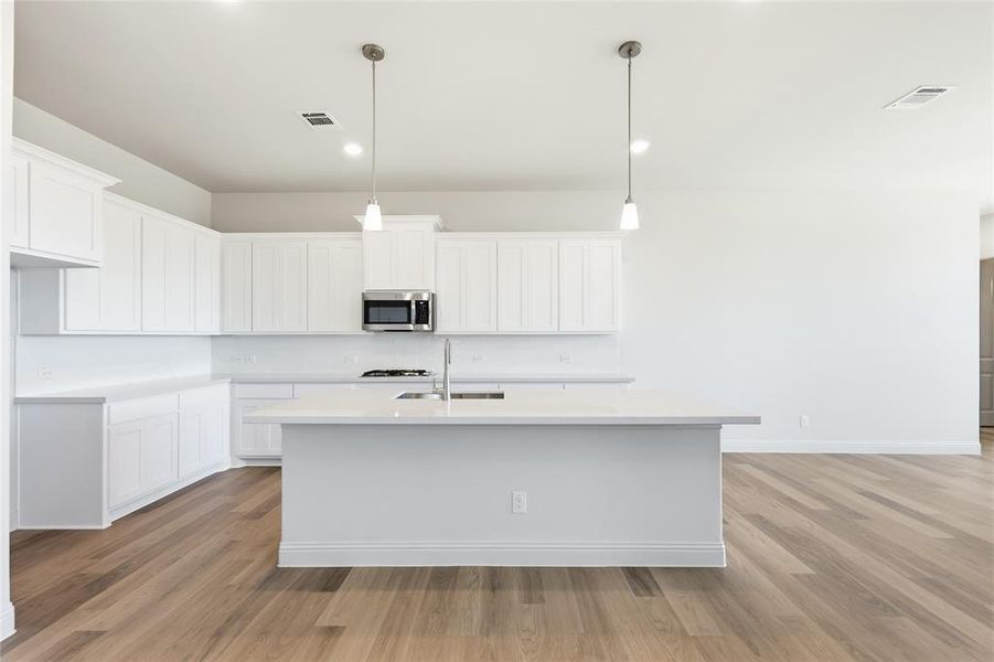 Kitchen with white cabinetry, a kitchen island with sink, light wood-type flooring, stainless steel microwave, and hanging light fixtures