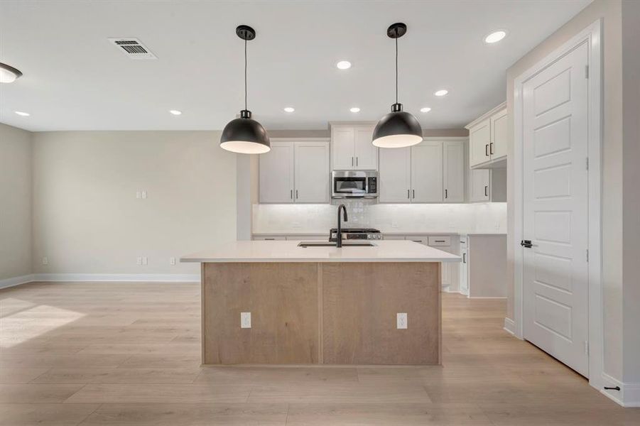 Two tone kitchen with a center island with sink, tasteful backsplash, stainless steel microwave, decorative light fixtures, and light wood-style floors