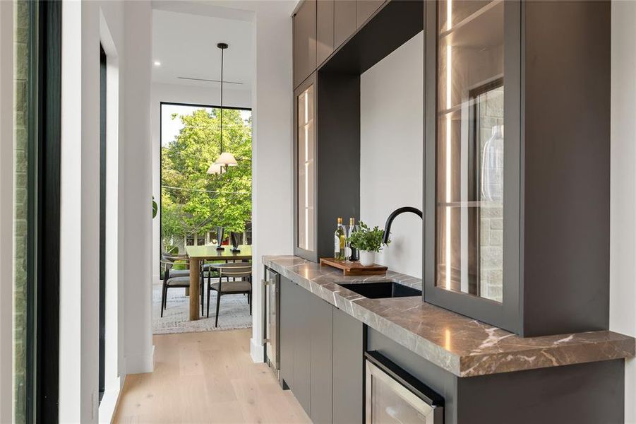 Built-in wet bar featuring dark cabinetry, stone countertop, integrated sink, and glass-front upper cabinets with interior lighting Built-in wet bar featuring dark cabinetry, stone countertop, integrated sink, and glass-front upper cabinets with interior lighting