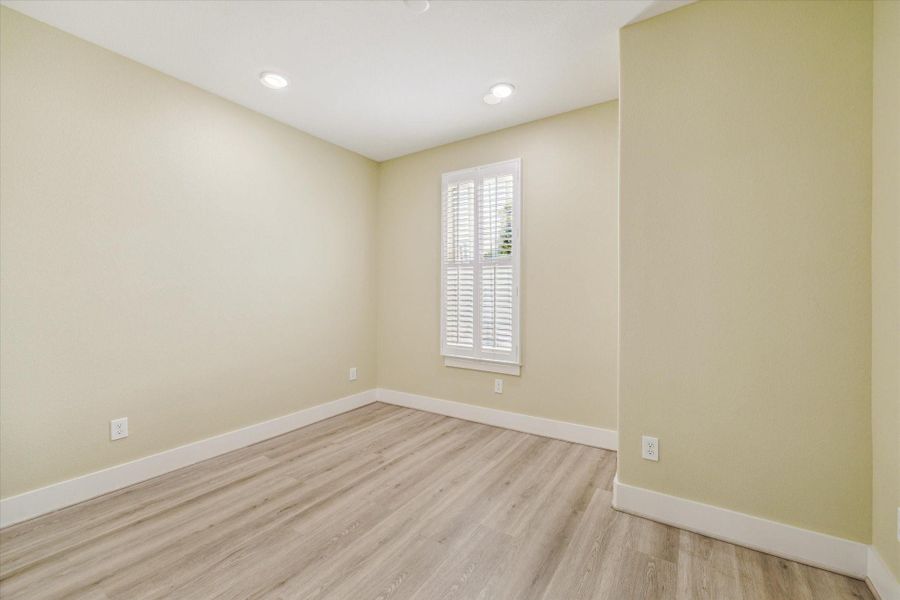 Bedroom 2 of 3,  This photo shows a bright, unfurnished room with light wood flooring and soft beige walls. It features a window with white shutters, recessed lighting, and clean baseboards, creating a modern and inviting space.