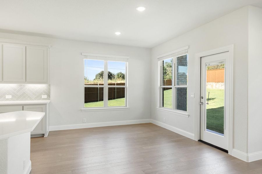 Bright dining area with light wood floors, two windows, and glass back door overlooking fenced backyard