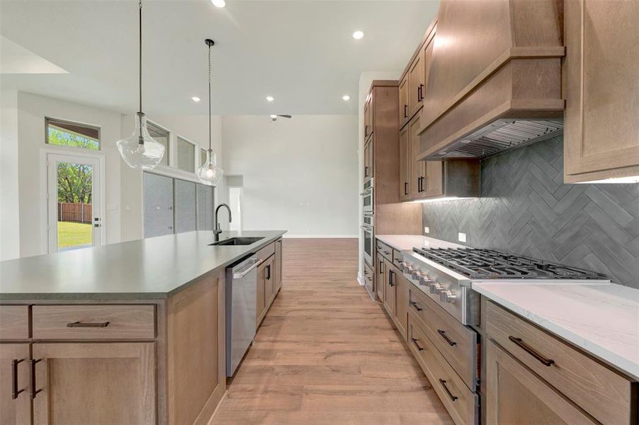 Kitchen with light wood-type flooring, stainless steel appliances, backsplash, light stone countertops, and a large island