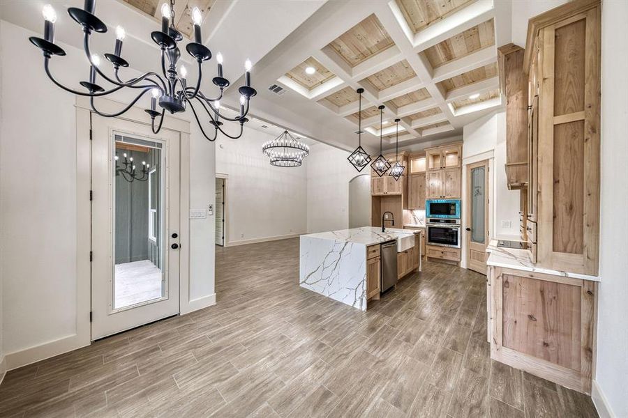 Kitchen featuring a kitchen island with sink, coffered ceiling, light stone countertops, wood finish floors, and stainless steel appliances