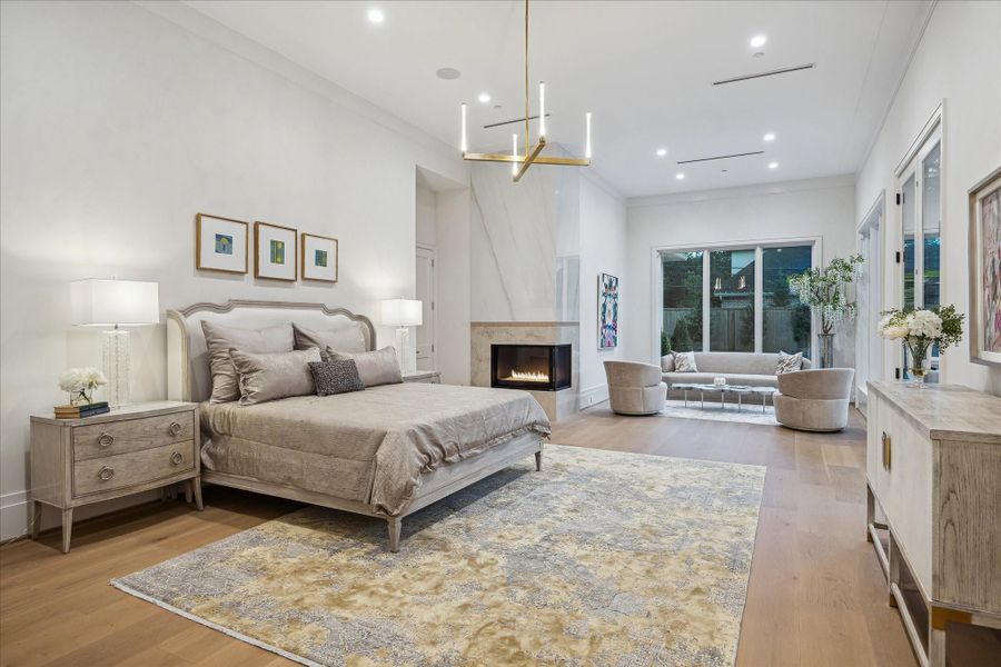 Serene luxurious primary suite featuring sitting space, designer chandelier, and a sleek gas corner fireplace with stone surround.