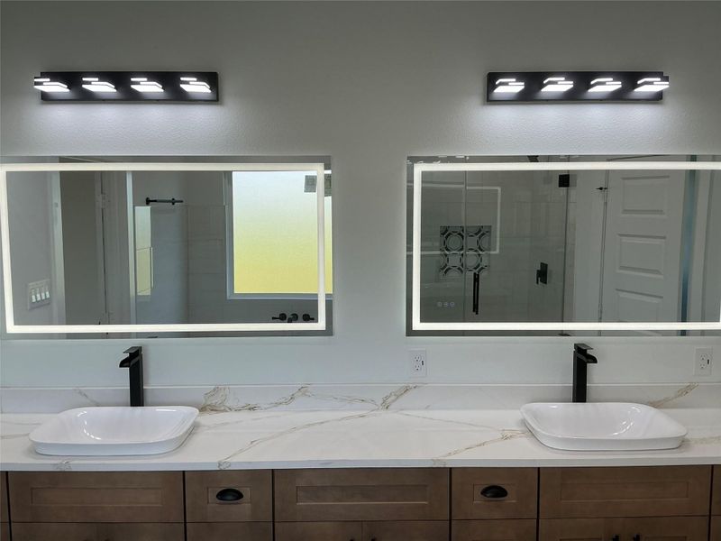 The master bathroom features a modern double vanity with sleek black fixtures, illuminated mirrors, and a quartz countertop. The space is well-lit with contemporary lighting and offers a clean, elegant design.