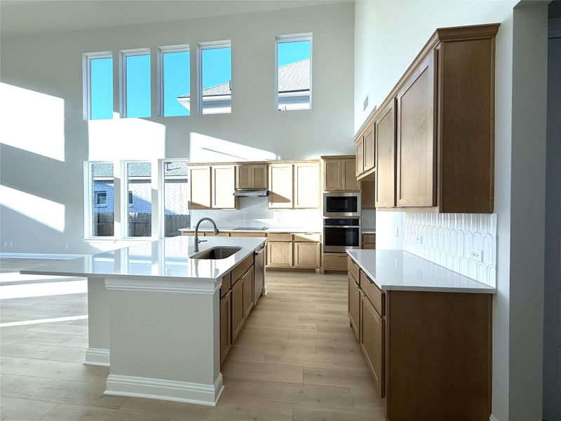 Kitchen with decorative backsplash, light stone counters, a kitchen island with sink, appliances with stainless steel finishes, and a towering ceiling