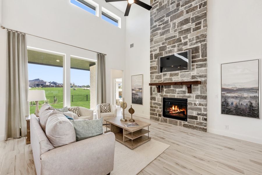Living room with two-story stone fireplace, ceiling fan, large windows, and light hardwood floors