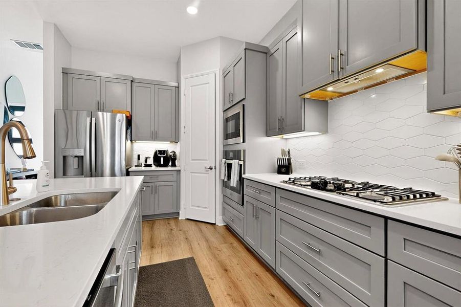 Kitchen featuring gray cabinetry, appliances with stainless steel finishes, backsplash, light wood finished floors, and light stone countertops Kitchen featuring gray cabinetry, appliances with stainless steel finishes, backsplash, light wood finished floors, and light stone countertops