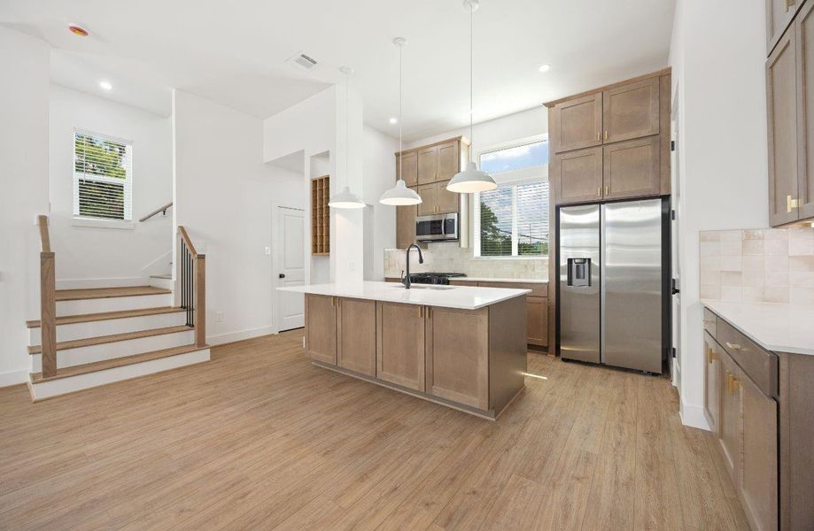 This open kitchen combines style and function with a large island, pendant lighting, warm wood cabinetry, quartz countertops, stainless steel refrigerator, gas cooking, and ample cabinet storage. The nearby staircase and open floor plan keep the kitchen connected to the main living spaces for easy entertaining.
