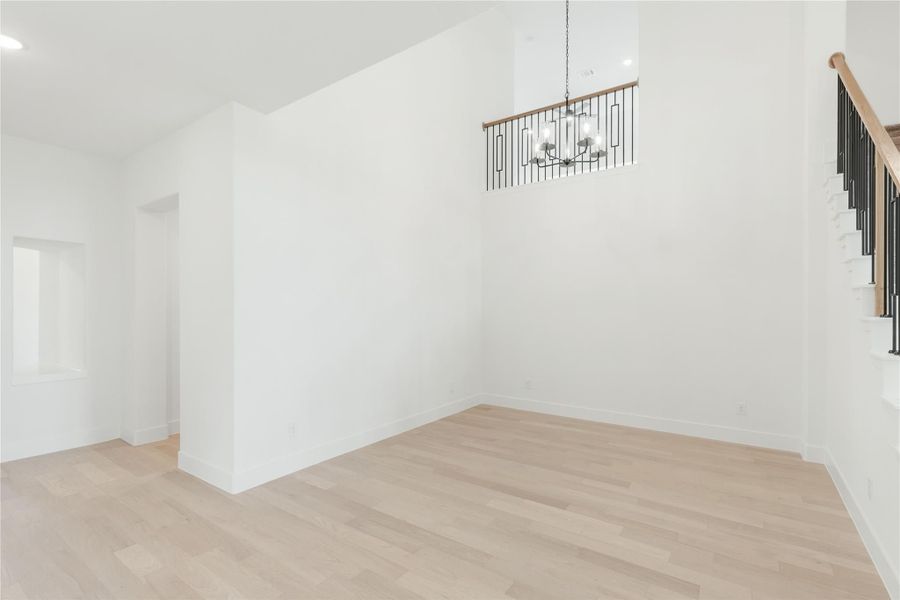 Empty dining area with high ceilings, light hardwood floors, chandelier, and staircase with black metal railing nearby.