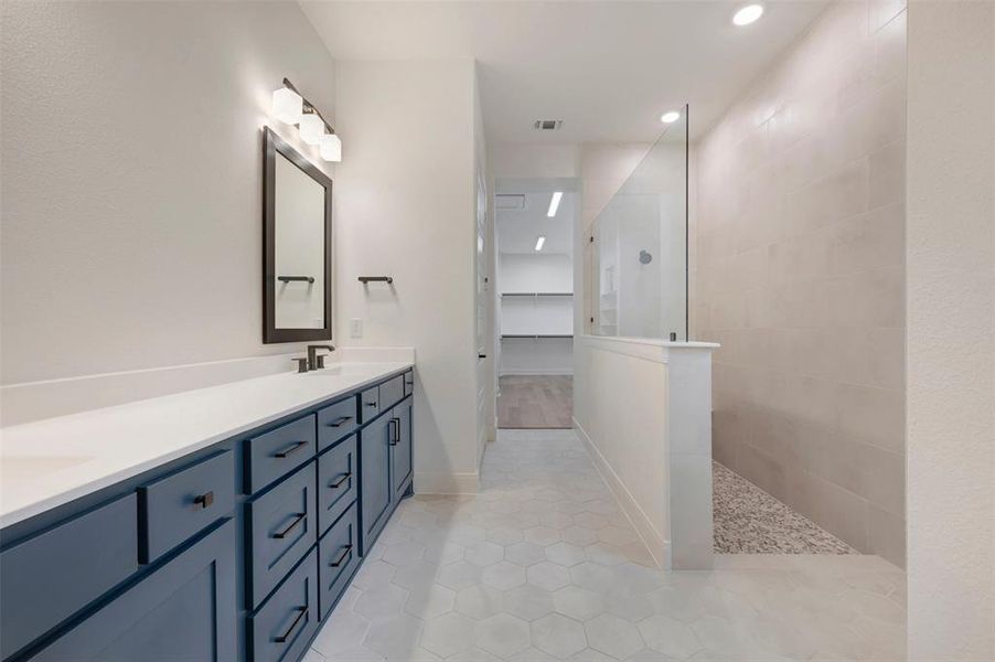 Bathroom featuring hexagon tiled floors, a long vanity with blue cabinetry and white countertops, a mirror with sconce lighting, and a glass-enclosed shower with pebble flooring