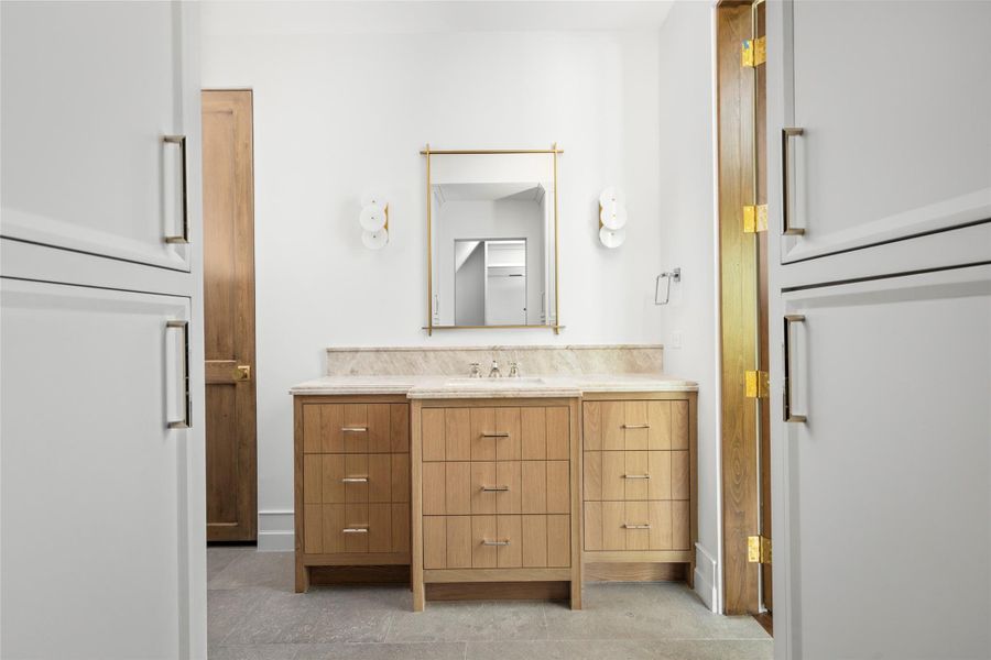 A secondary bath pairs a warm wood vanity with a softly veined natural stone countertop and backsplash. Floor-to-ceiling built-in cabinetry provides generous storage, while brass accents and sculptural sconces give the space a polished, tailored finish. A secondary bath pairs a warm wood vanity with a softly veined natural stone countertop and backsplash. Floor-to-ceiling built-in cabinetry provides generous storage, while brass accents and sculptural sconces give the space a polished, tailored finish.