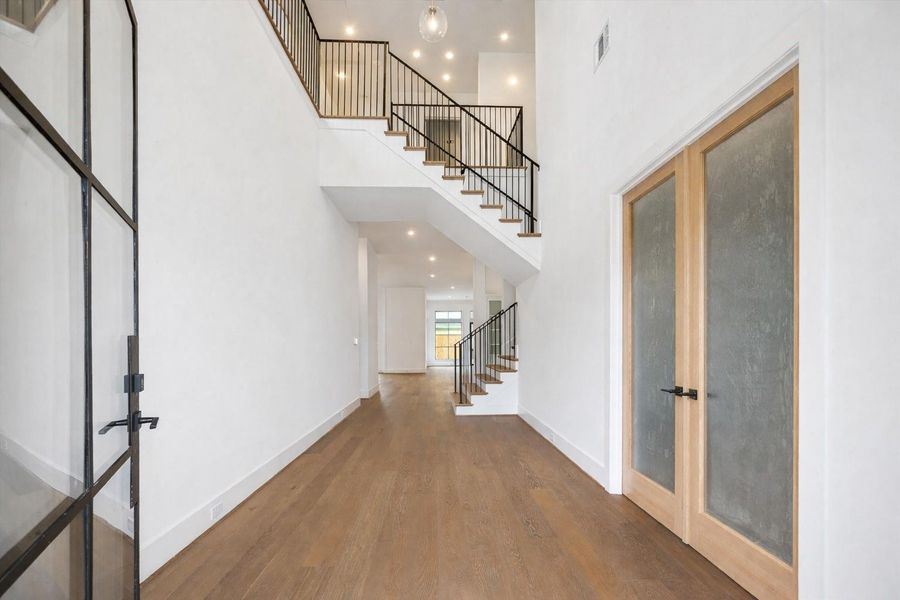 Soaring two story entry with wide plank hardwood floors and a modern iron staircase creates a dramatic first impression. Glass front door, clean white walls, and warm wood accents offer seamless flow into the open concept living spaces beyond, blending contemporary design with timeless architectural detail.