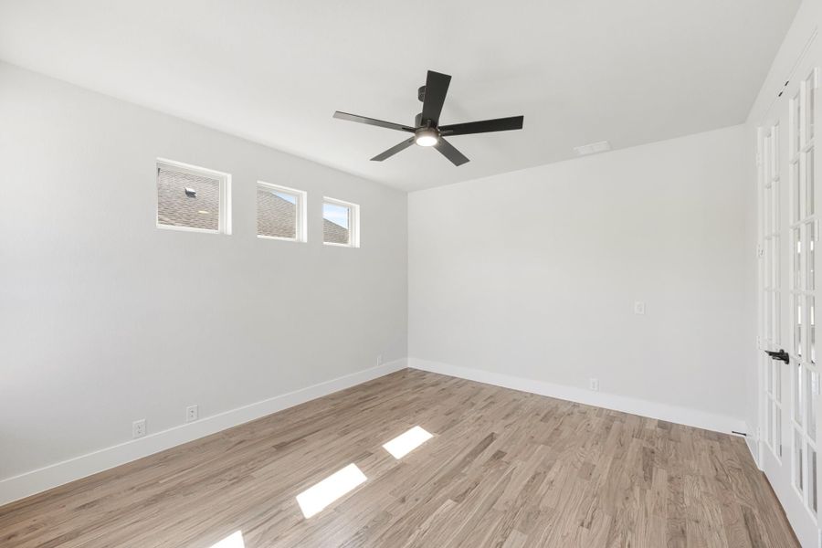 Empty bedroom with light wood floors, white walls, black ceiling fan, and small windows