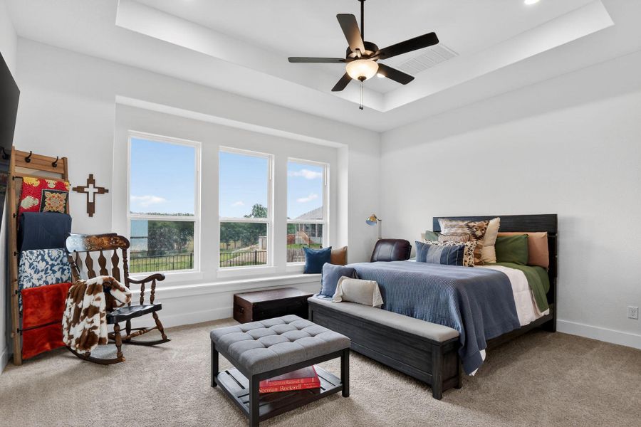 Bedroom featuring a tray ceiling with a ceiling fan, three large windows with white trim, and light-colored carpet flooring Bedroom featuring a tray ceiling with a ceiling fan, three large windows with white trim, and light-colored carpet flooring