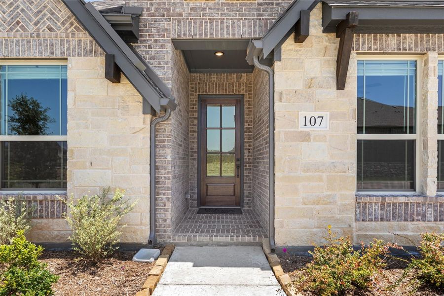 Front entry of home number 107 with brick and stone exterior, covered porch, and wood door with glass panel Front entry of home number 107 with brick and stone exterior, covered porch, and wood door with glass panel