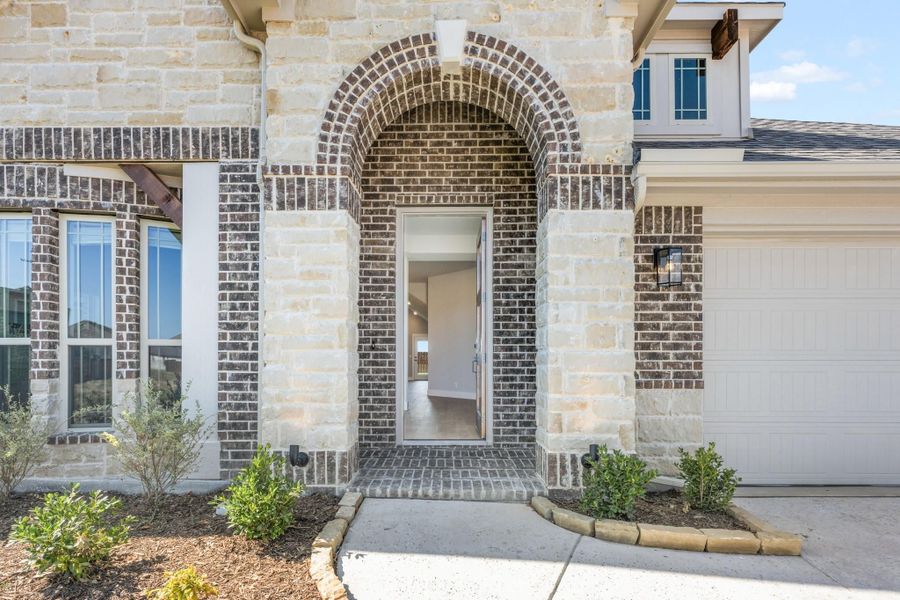 Home entrance with arched brick doorway, stone exterior, and white garage door with landscaped shrubs nearby Home entrance with arched brick doorway, stone exterior, and white garage door with landscaped shrubs nearby