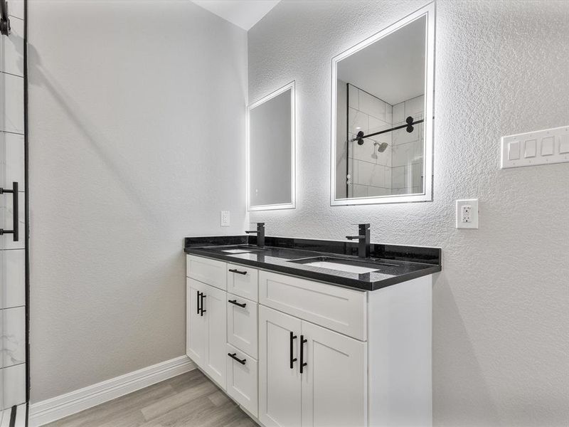 Bathroom with wood finished floors, a tile shower, double vanity, a textured wall, and baseboards