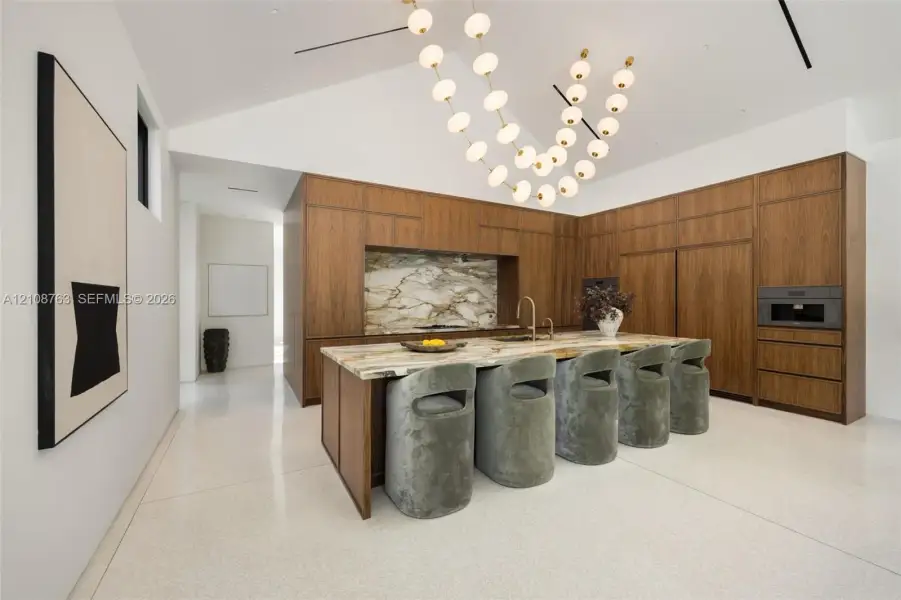 Modern kitchen featuring full-height wood cabinetry, a stone-top island with integrated sink, polished light-colored flooring, vaulted ceiling, and tiered orb pendant lighting