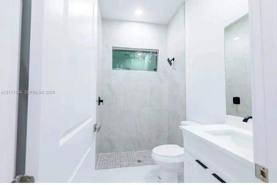Bathroom featuring a shower stall with full-height tile surrounds, a transom window, matte black hardware, and a white vanity with an integrated sink Bathroom featuring a shower stall with full-height tile surrounds, a transom window, matte black hardware, and a white vanity with an integrated sink
