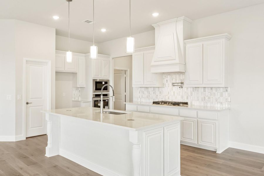 White kitchen with large island, pendant lights, custom range hood, and tile backsplash on hardwood floors