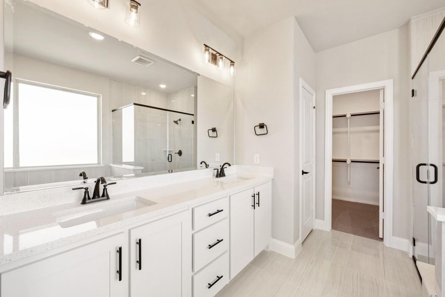 Master bathroom with double vanity, white cabinets, quartz countertops, and walk-in closet entrance visible. Master bathroom with double vanity, white cabinets, quartz countertops, and walk-in closet entrance visible.