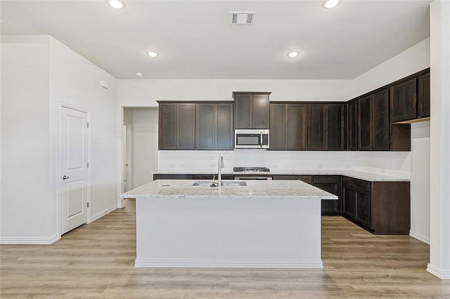 Kitchen with sink, an island with sink, dark brown cabinets, and light wood-type flooring