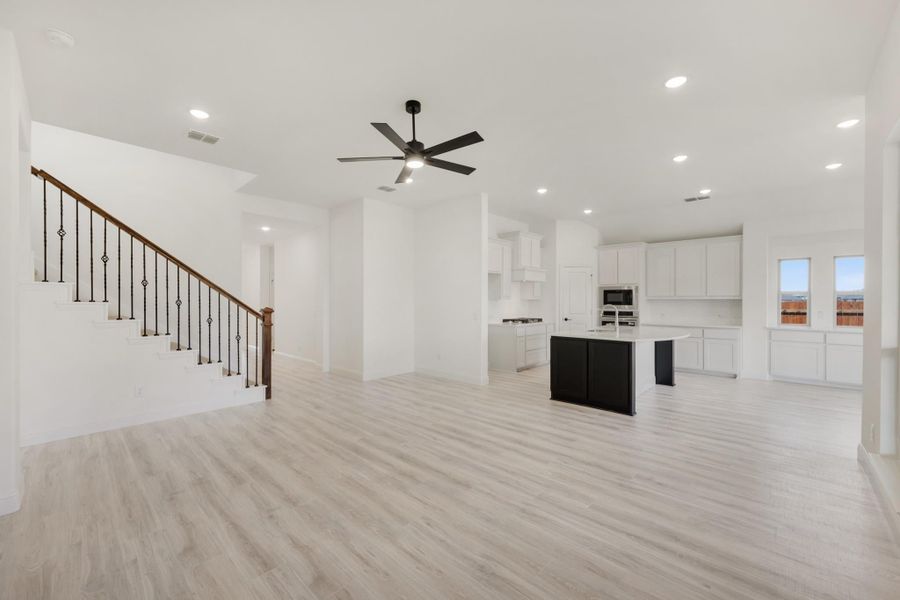 Open living area with light wood floors, ceiling fan, staircase, and kitchen with dark island and white cabinets