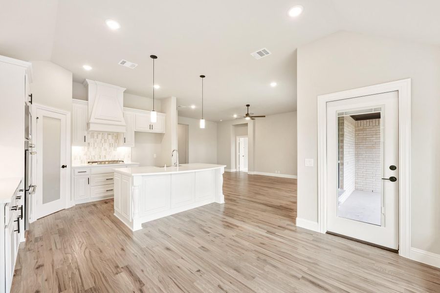 Open kitchen with white cabinets, large island, pendant lights, and light wood floors opening to living area Open kitchen with white cabinets, large island, pendant lights, and light wood floors opening to living area