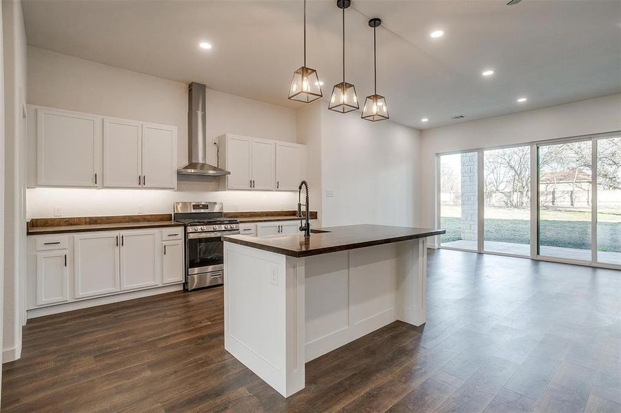 Kitchen with stainless steel gas range, wall chimney exhaust hood, pendant lighting, an island with sink, and white cabinetry