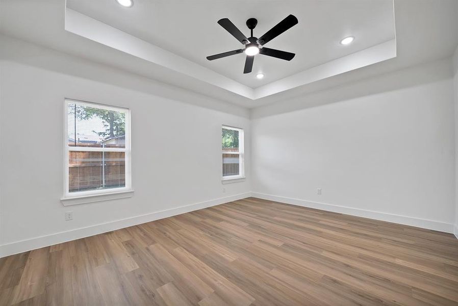 Spacious room featuring wood-finish flooring, two windows with white trim, a tray ceiling, recessed lighting, and a black ceiling fan Spacious room featuring wood-finish flooring, two windows with white trim, a tray ceiling, recessed lighting, and a black ceiling fan