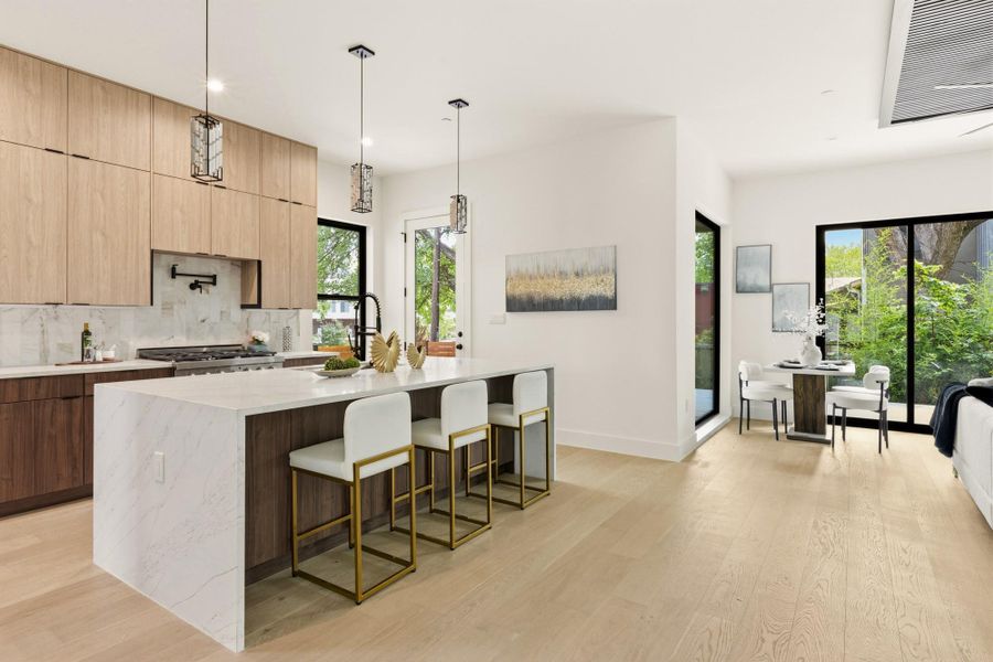 Open-concept kitchen and dining area featuring a waterfall island with a light-toned countertop, wood-finish cabinetry, and light wood-finish flooring