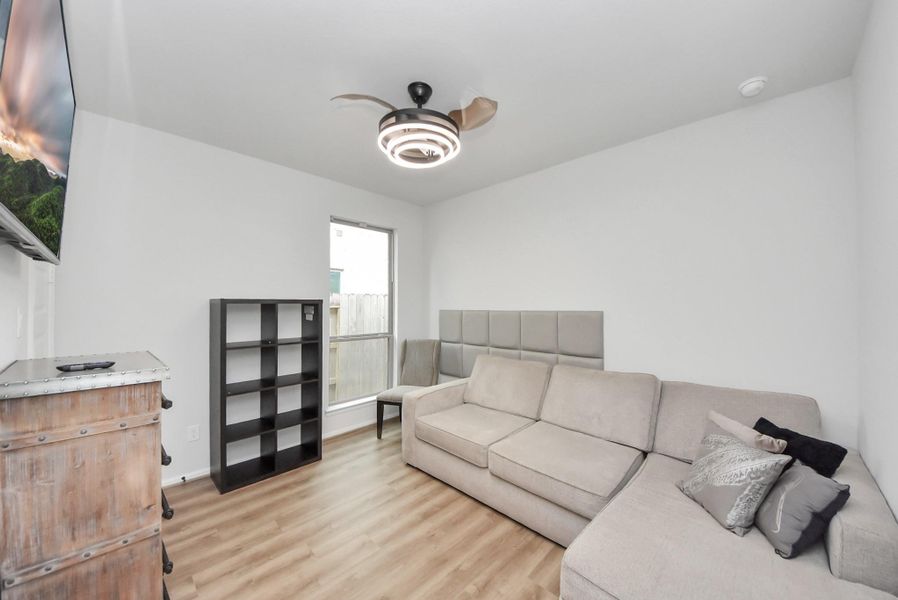 This cozy Bedroom features a modern ceiling fan, light wood flooring, and a large sectional sofa. There's a wall-mounted TV, a sleek shelving unit, and a window providing natural light, creating a comfortable and inviting space.