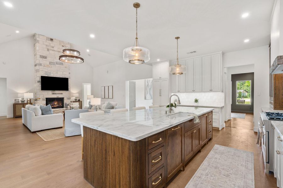 Showpiece kitchen featuring a fully paneled refrigerator that blends seamlessly into the custom cabinetry for a clean, built-in look. The oversized island offers substantial prep space and storage, anchored by upgraded pendant lighting overhead. A professional-style range with pot filler sits beneath a detailed vent hood, framed by warm wood cabinetry for contrast. Floor-to-ceiling cabinets, extensive drawer storage, and thoughtful layout design to truly give a "wow" factor!