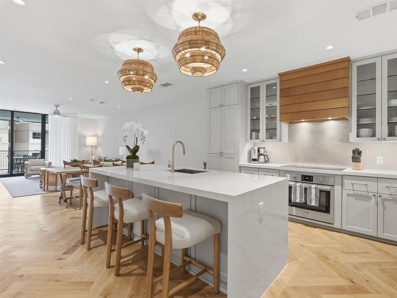 Open-concept kitchen featuring a waterfall island with integrated seating, a wood-finish range hood, stainless steel appliances, and herringbone wood-finish flooring Open-concept kitchen featuring a waterfall island with integrated seating, a wood-finish range hood, stainless steel appliances, and herringbone wood-finish flooring