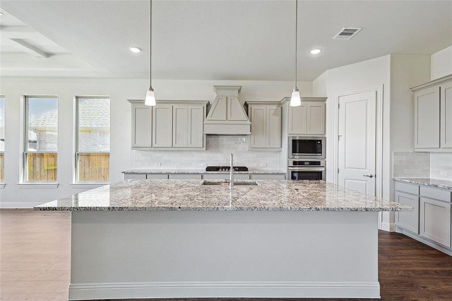 Kitchen with dark wood-type flooring, tasteful backsplash, stainless steel appliances, a center island with sink, and light stone countertops