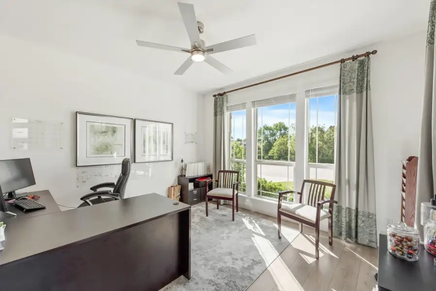 Home office with dark wood desk, ceiling fan, hardwood floors, and large windows with natural light