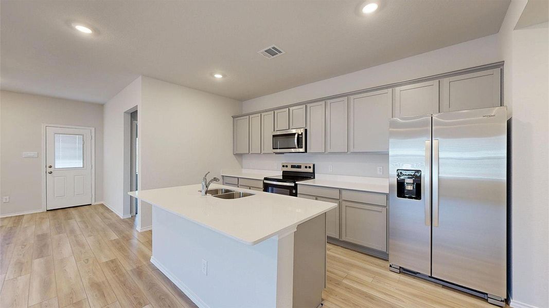 Modern kitchen featuring an island with an undermount double-basin sink, light-toned countertops, light gray cabinetry, stainless steel appliances, and wood-finish flooring
