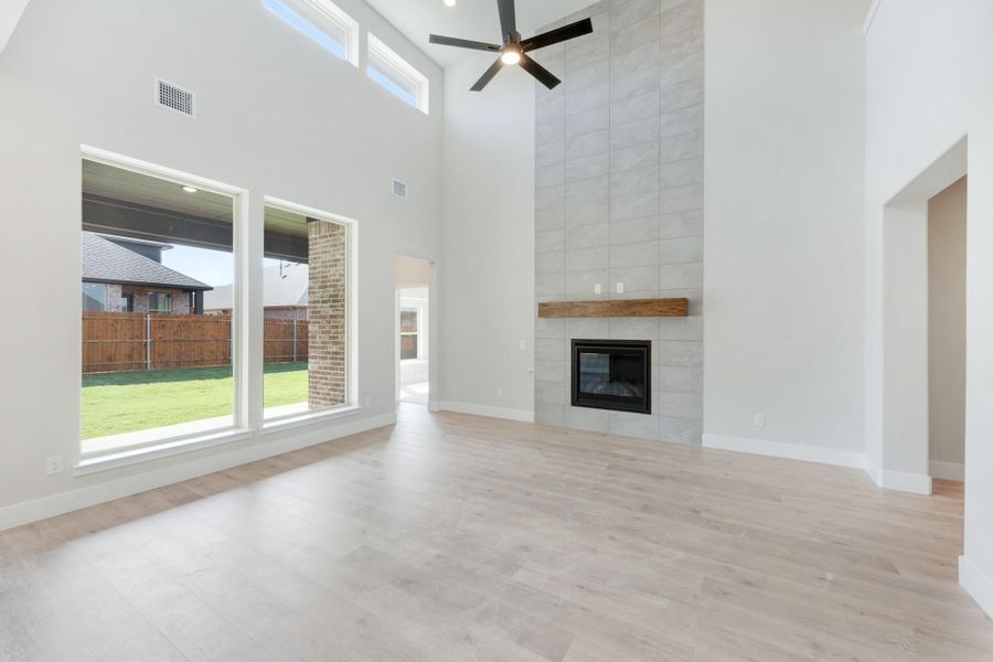 Open living room with two-story ceiling, tile fireplace, ceiling fan, and large windows overlooking backyard