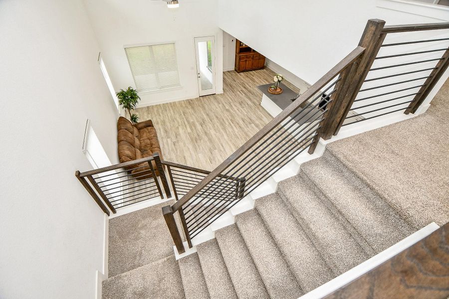 This photo highlights a modern, open-concept living area with carpeted stairs descending to a bright, welcoming space. A large window allows plenty of natural light, complemented by a stylish horizontal iron railing that adds a contemporary touch. This photo highlights a modern, open-concept living area with carpeted stairs descending to a bright, welcoming space. A large window allows plenty of natural light, complemented by a stylish horizontal iron railing that adds a contemporary touch.