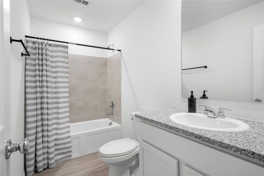 Full bathroom featuring a white vanity with a speckled countertop, an undermount sink with chrome fixtures, a built-in bathtub with neutral-toned tile surround, and wood-finish flooring Full bathroom featuring a white vanity with a speckled countertop, an undermount sink with chrome fixtures, a built-in bathtub with neutral-toned tile surround, and wood-finish flooring