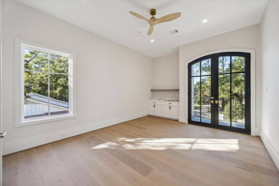 Bright study/exercise room featuring wide-plank wood floors, a built-in workstation with storage, and an elegant arched French door that opens onto a private balcony. Expansive windows bring in abundant natural light, creating a versatile space ideal for work, wellness, or relaxation.