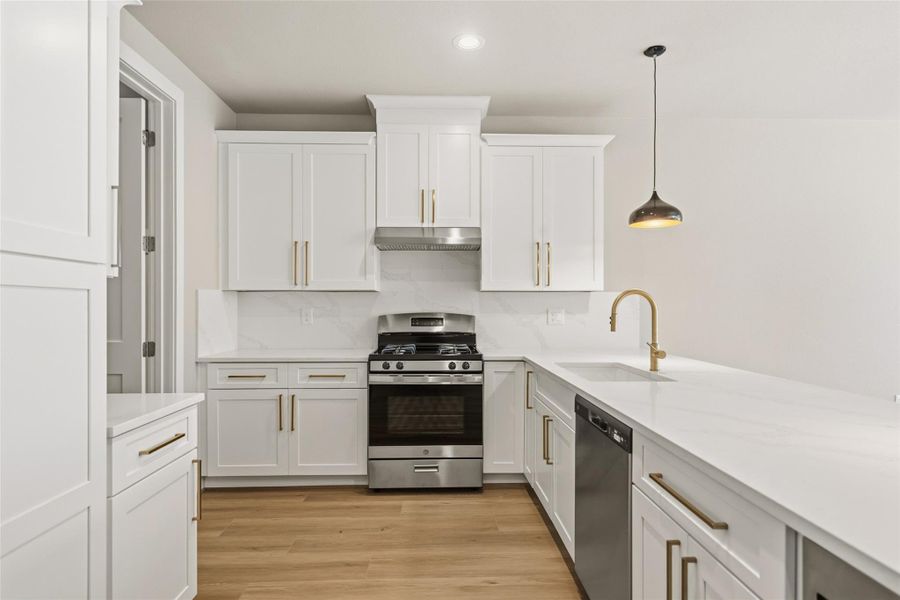This modern kitchen features sleek white cabinetry with gold handles, a stainless steel gas stove and dishwasher, and elegant quartz countertops. A stylish pendant light and wooden flooring add warmth and sophistication.