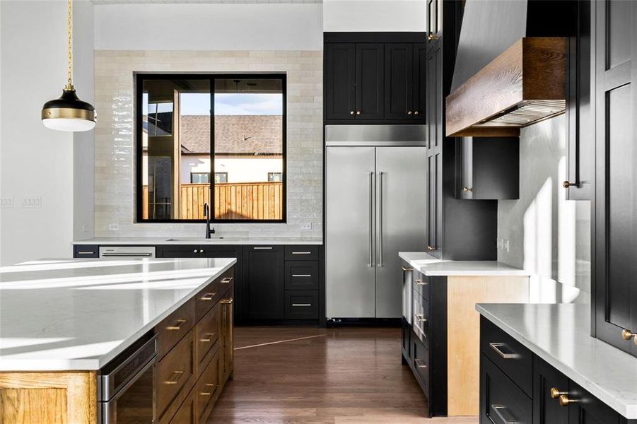 Kitchen with dark cabinets, stainless steel built in fridge, light stone counters, and premium range hood Kitchen with dark cabinets, stainless steel built in fridge, light stone counters, and premium range hood