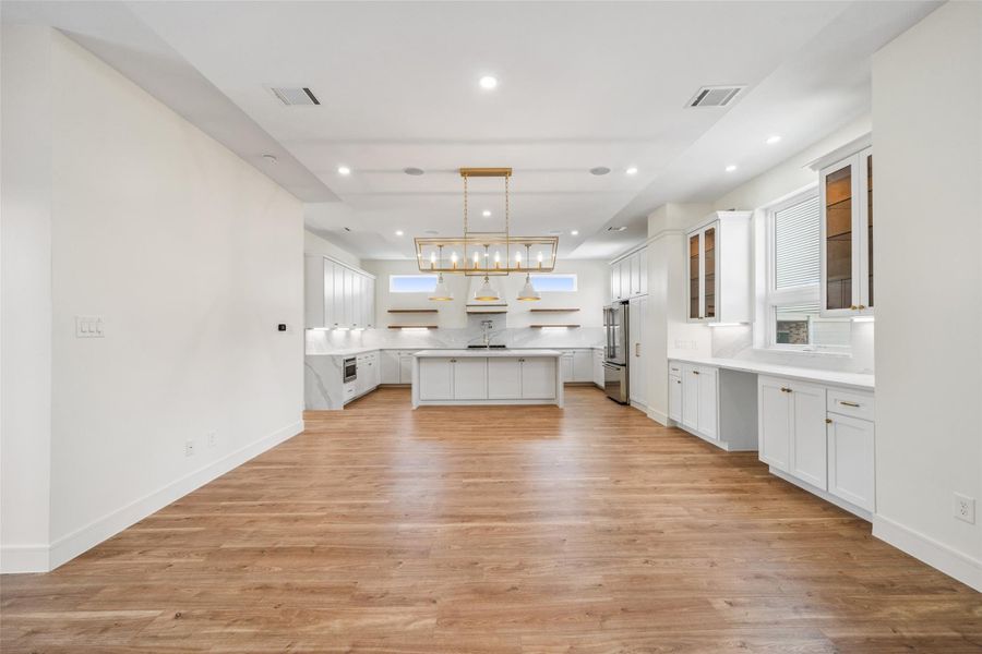 Open-concept kitchen and dining area with wood floors. The kitchen features white cabinetry, modern stainless steel appliances, and elegant pendant lighting over a large island. Bright, recessed lighting and ample windows provide a well-lit, inviting space.