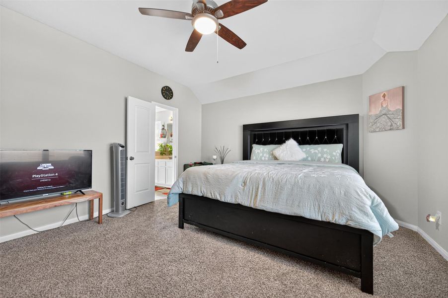 This cozy bedroom features a large bed with a modern headboard, neutral walls, and carpeted flooring. A ceiling fan provides comfort, while a TV setup adds entertainment options. An open door leads to an adjoining space, enhancing accessibility.