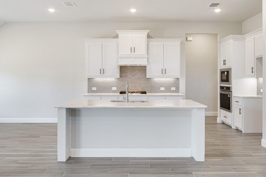 White kitchen with large center island, white cabinets, tile backsplash, and double wall ovens on light wood floors White kitchen with large center island, white cabinets, tile backsplash, and double wall ovens on light wood floors