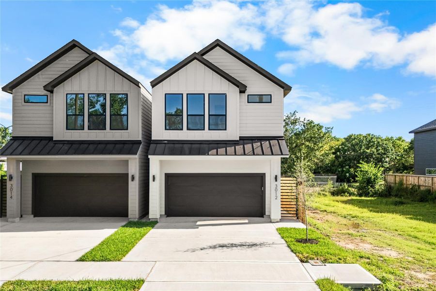 The contemporary exterior features a mix of light gray board-and-batten and horizontal lap siding on the upper level, with smooth light gray stucco below. A black metal awning separates the levels, framing the massive windows that bring in natural light.