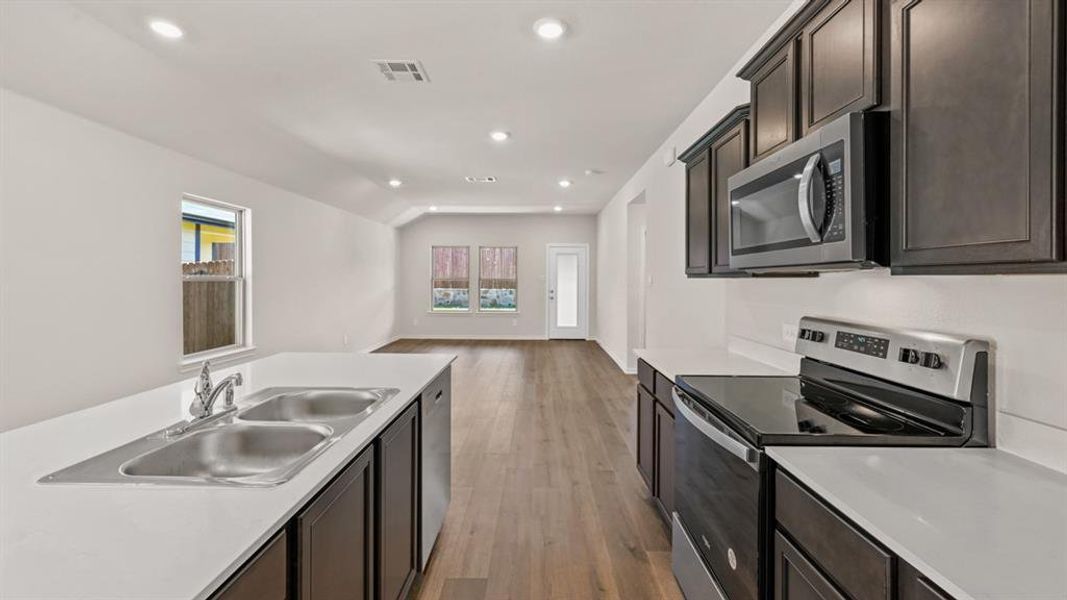 Open-concept kitchen featuring an island with a double stainless steel sink, dark wood cabinetry, white countertops, and stainless steel appliances, including an electric range and microwave, all complemented by wood-finish flooring and recessed lighting